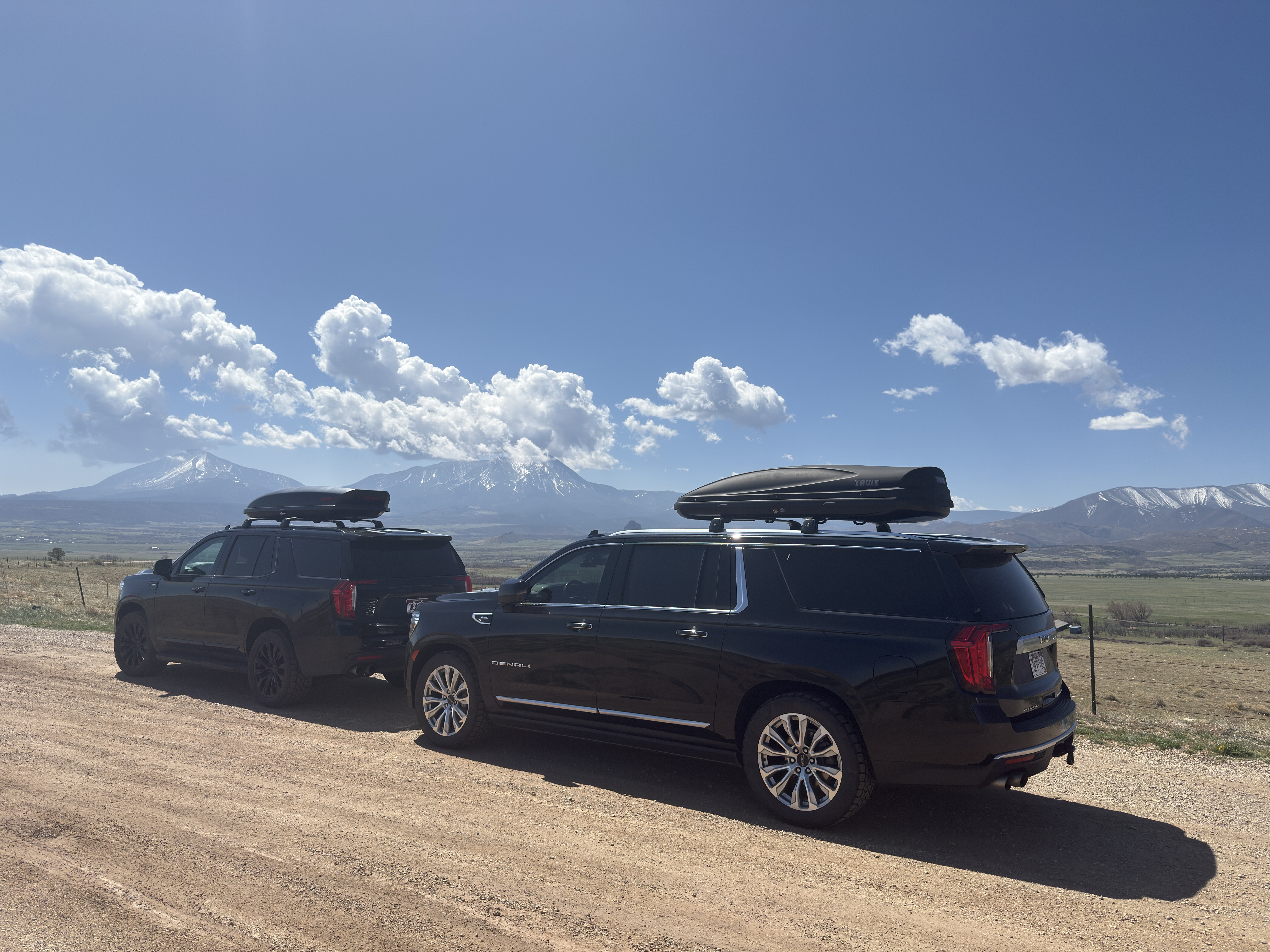 Two black GMC Yukon Denalis equipped with rooftop carriers, navigating a scenic dirt road with a vast panoramic mountain landscape and expansive blue sky.