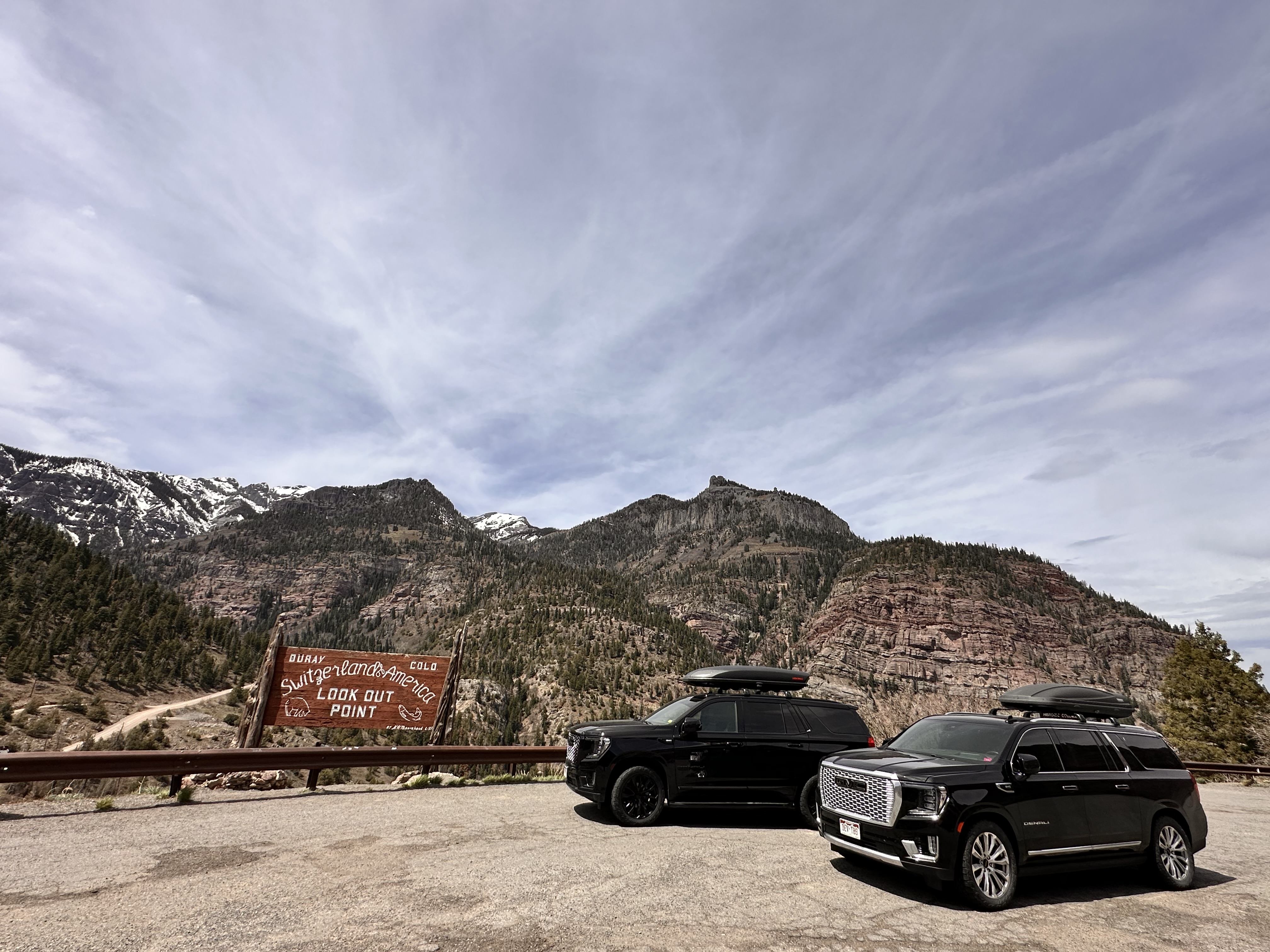 Two black GMC Yukon Denali SUVs parked side-by-side at the 'Switzerland of America' scenic lookout, with stunning snow-capped mountains under a clear blue sky.