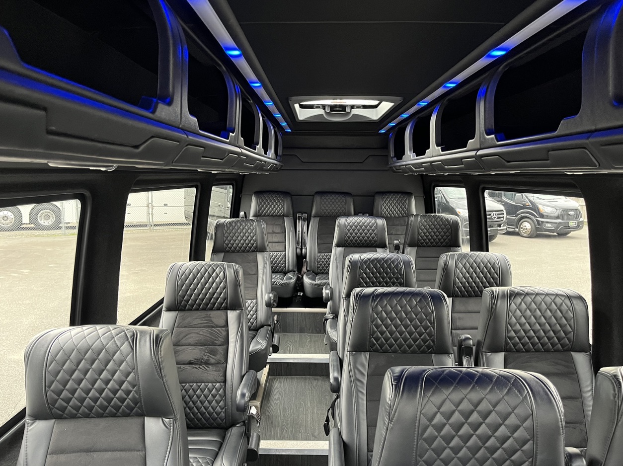 Interior view of a luxury Sprinter van showcasing multiple rows of black quilted leather seats and ambient blue LED accent lighting, emphasizing comfort and style.